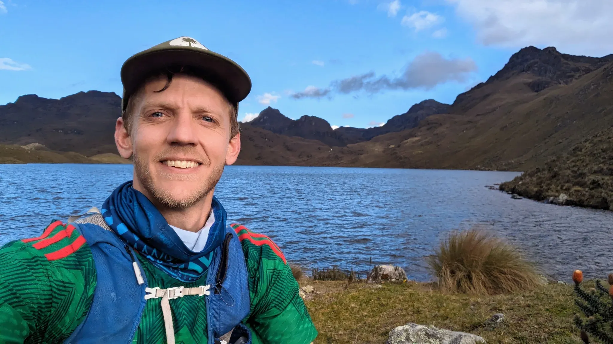 Travis wearing a cap and running vest beside a lake, with rocky mountains behind him.
