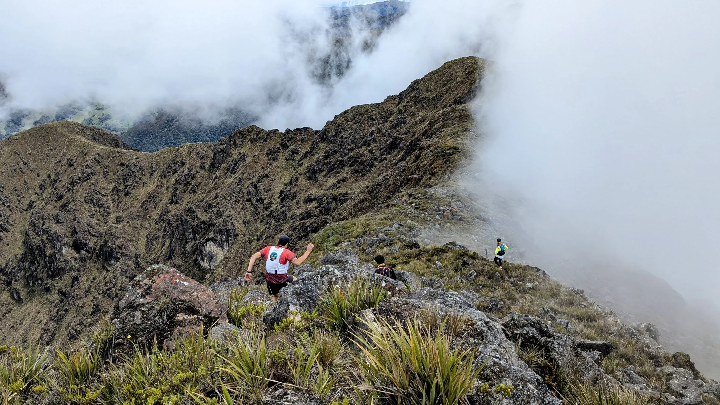 Tres corredores en una cresta rocosa con pajonal, con nubes que suben por la derecha.